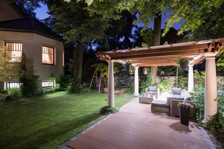 A backyard at night with a pergola over a patio illuminated by landscape lighting.