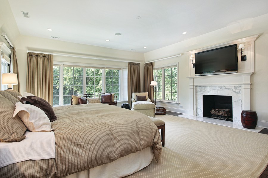 Expansive bedroom with a king size bed, a chair, a window bench, in-ceiling speakers, wall-mounted TV,  and view of the trees outside. 