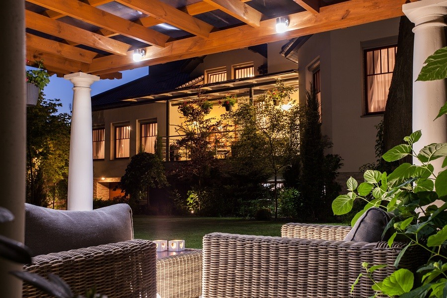 Beautifully illuminated home with landscape lighting as seen from under a pergola and wicker furniture.  