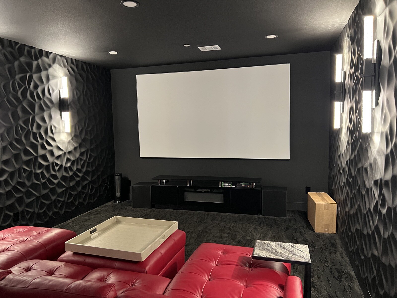 Angled view of a projection screen, red leather seating, and illuminated sconces along patterned theater walls