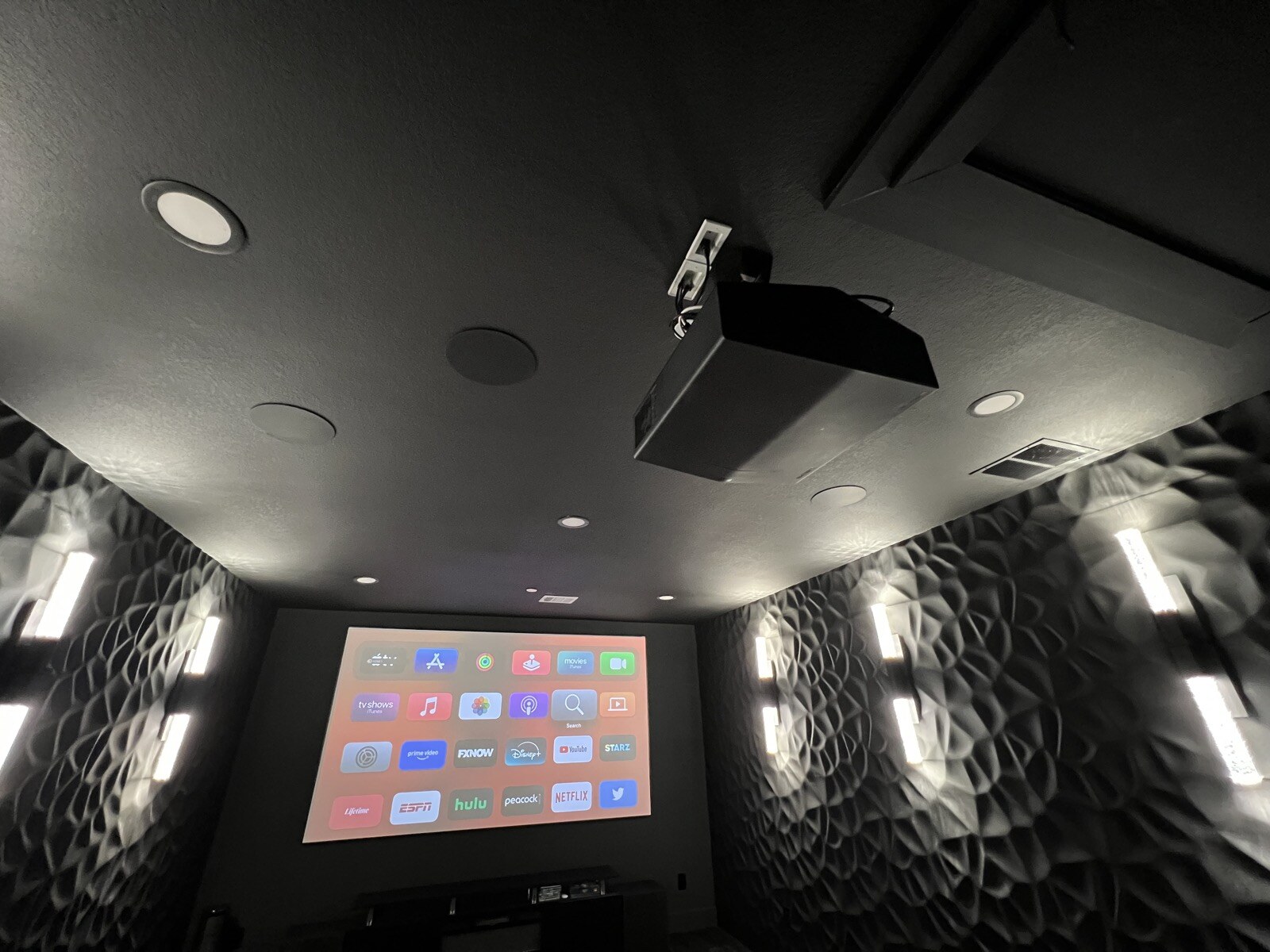 Ceiling-mounted projector and round ceiling speakers above a theater screen displaying streaming apps