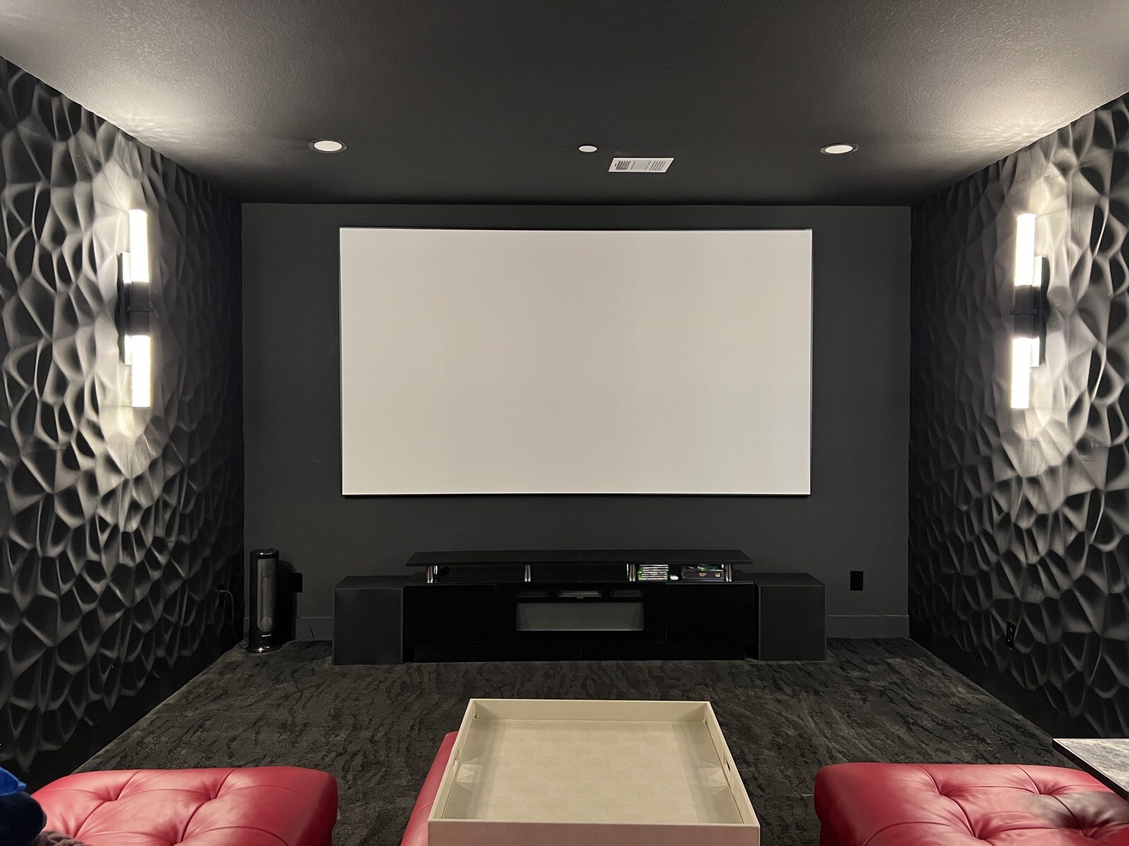 Home theater projection screen framed by patterned walls and illuminated sconces, with red leather seating in front
