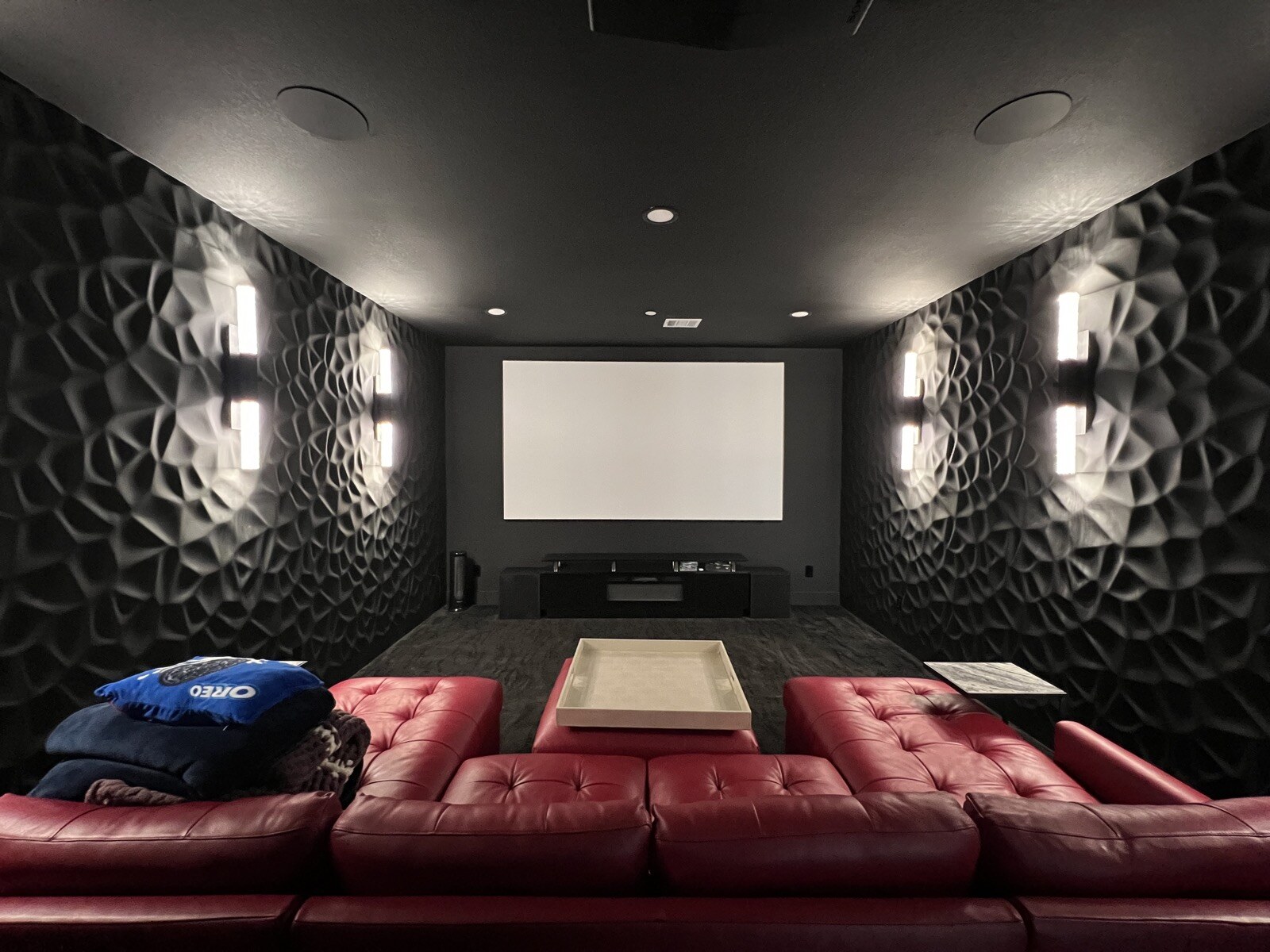 Illuminated wall sconces beside a home theater screen, with ceiling speakers and red leather seating