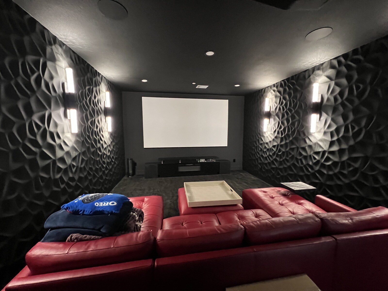Wide angled view over red leather seating toward a projection screen, with ceiling speakers and patterned walls