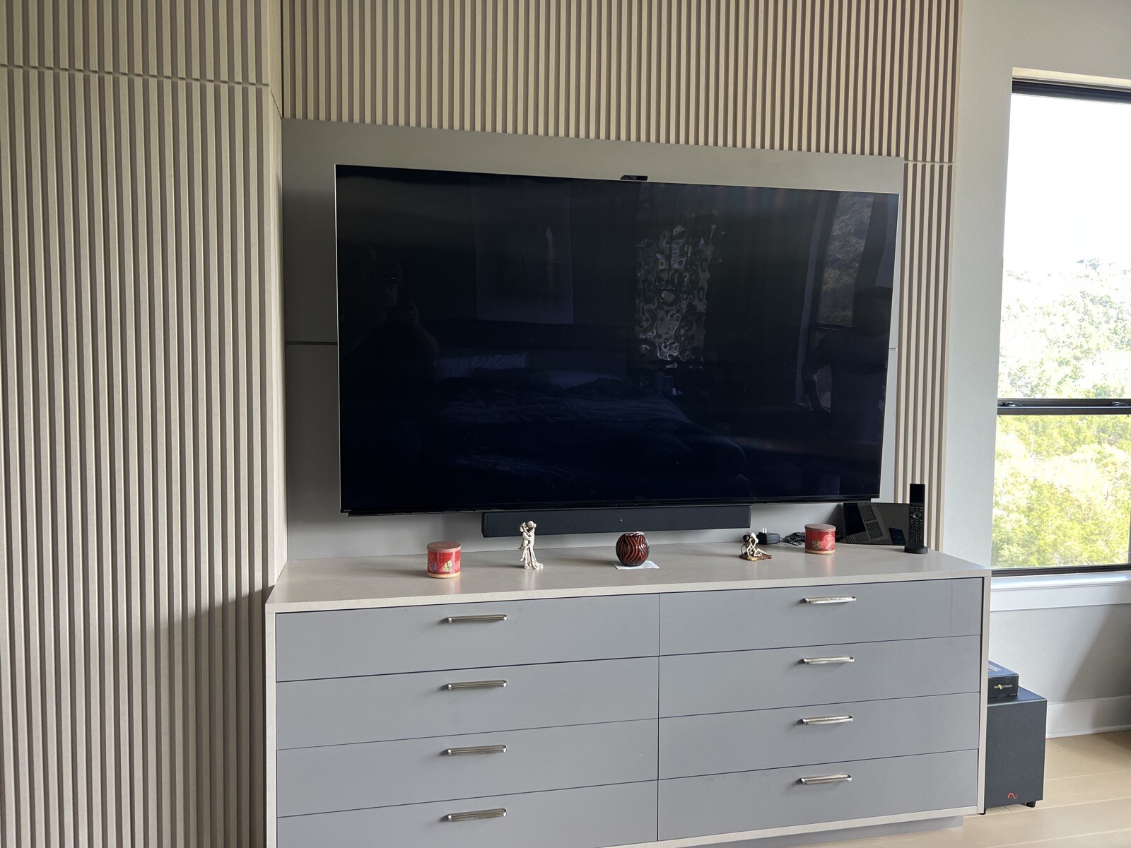 TV mounted on a vertical slat accent wall above white drawers beside a tall window