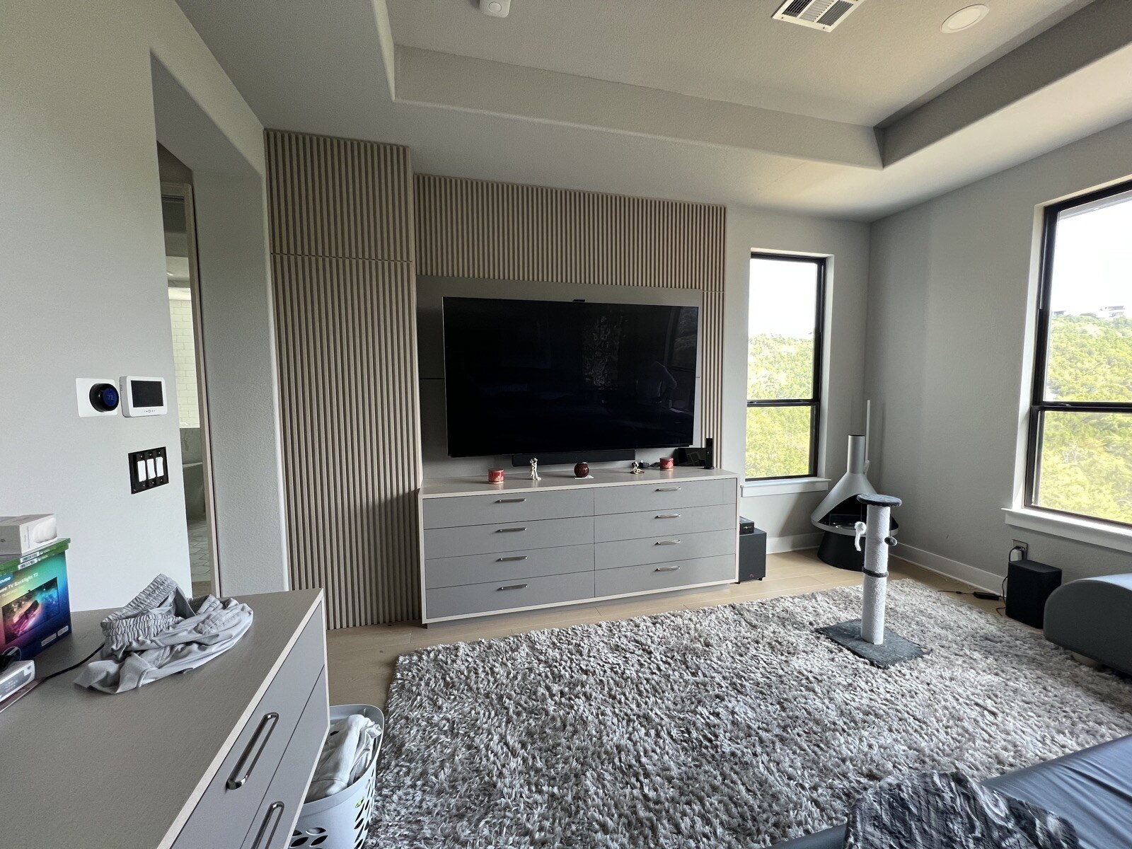 Wide view of a TV and white drawer cabinet against a slat wall, with tall windows and wall controls at left