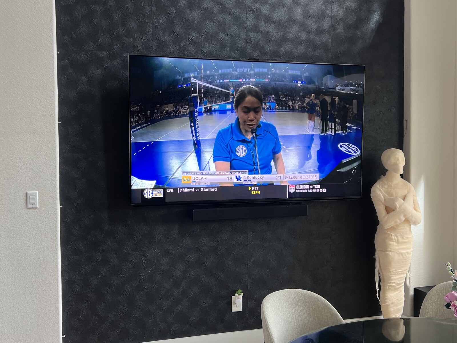 Wall-mounted TV showing a sports interview above a soundbar on dark patterned wallpaper