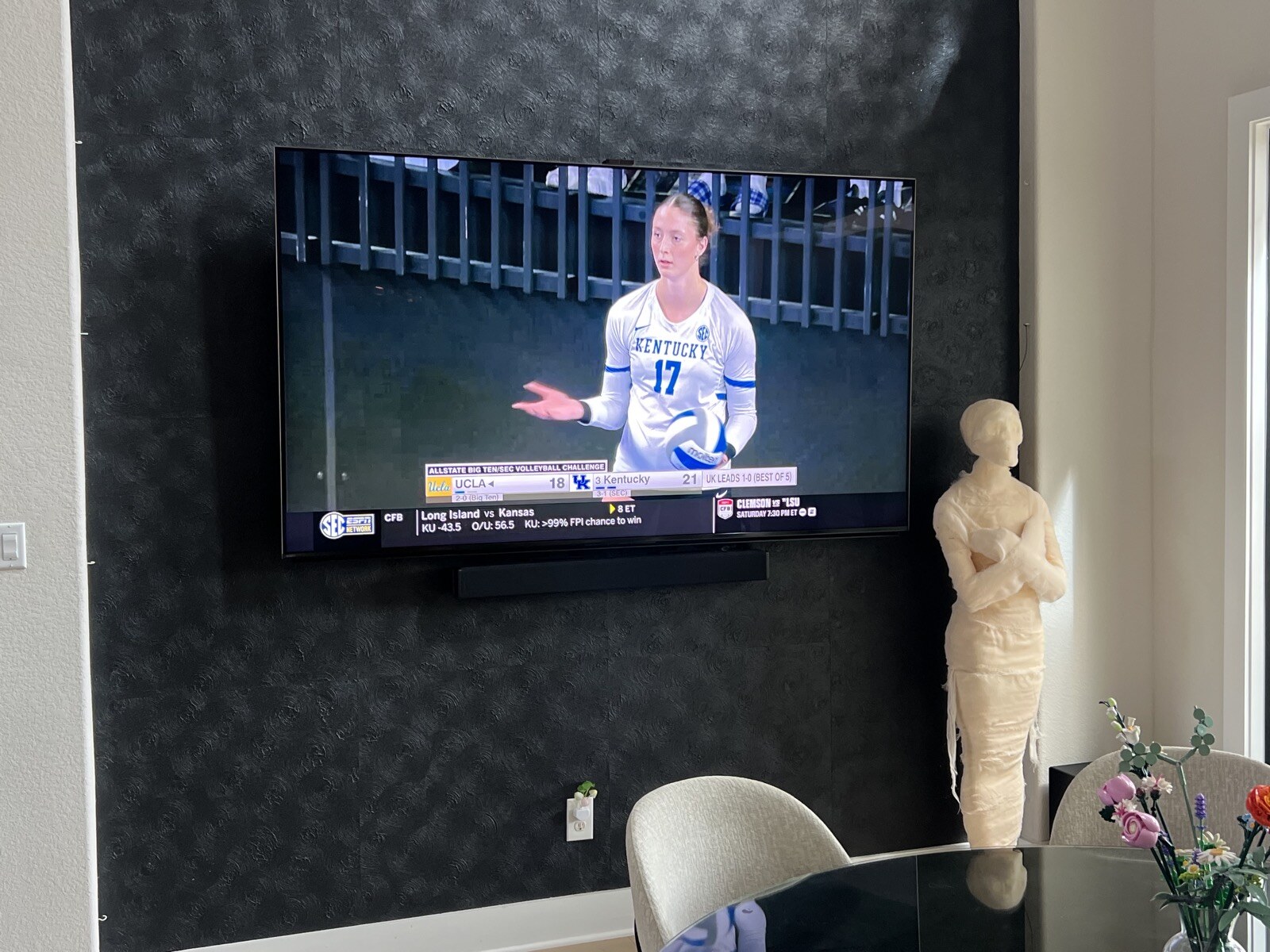 Volleyball broadcast on a wall-mounted TV above a soundbar, beside a decorative figure and table