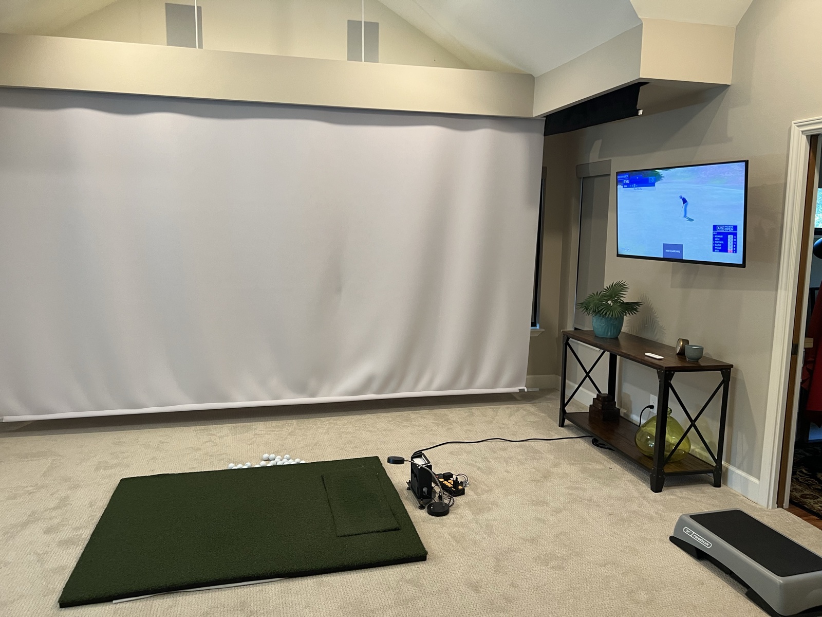 The golf simulator room with the impact screen down, a practice mat and balls on the floor, and live golf on the wall TV