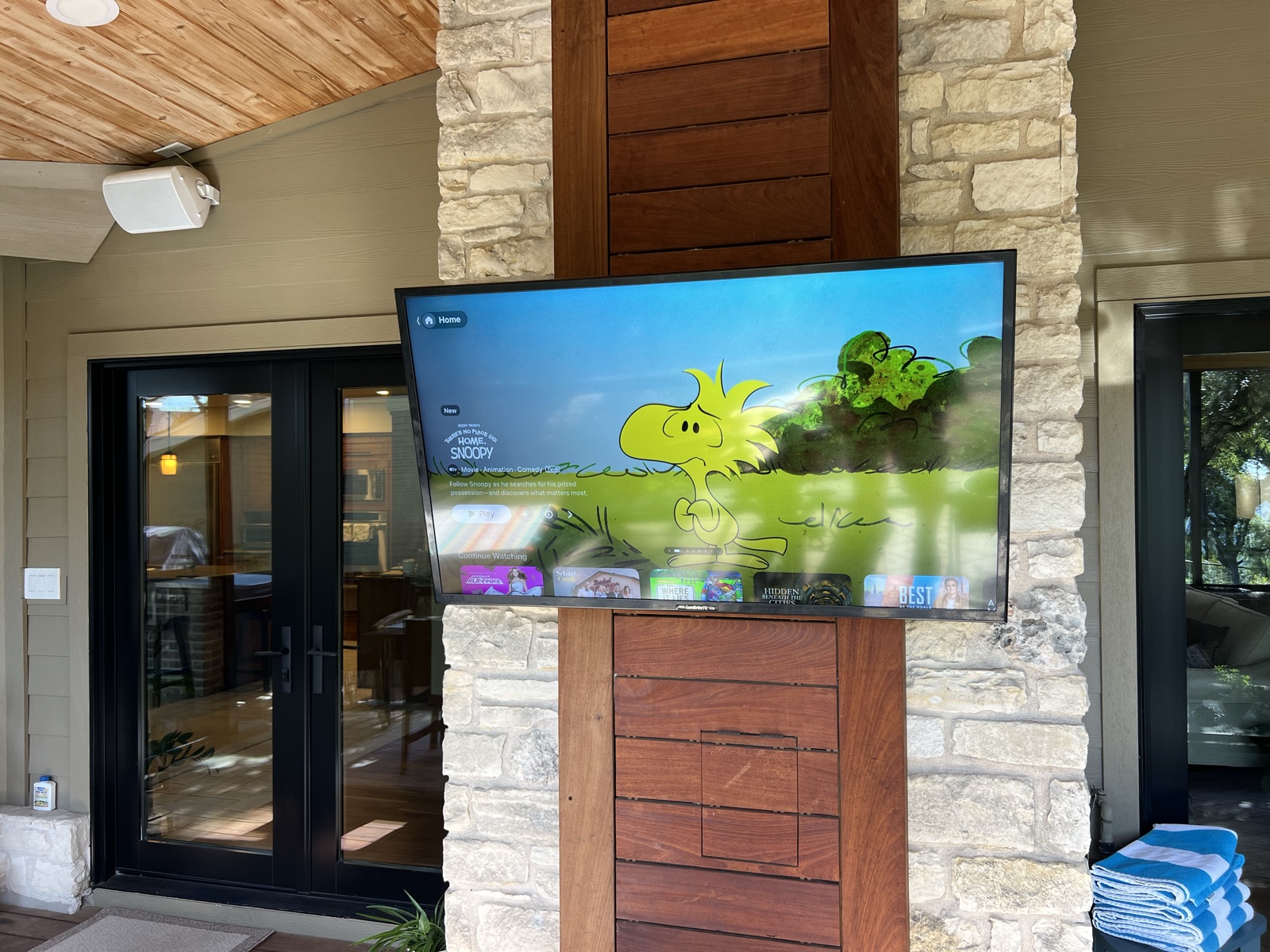 An outdoor TV playing on the stone patio column with an outdoor speaker mounted overhead