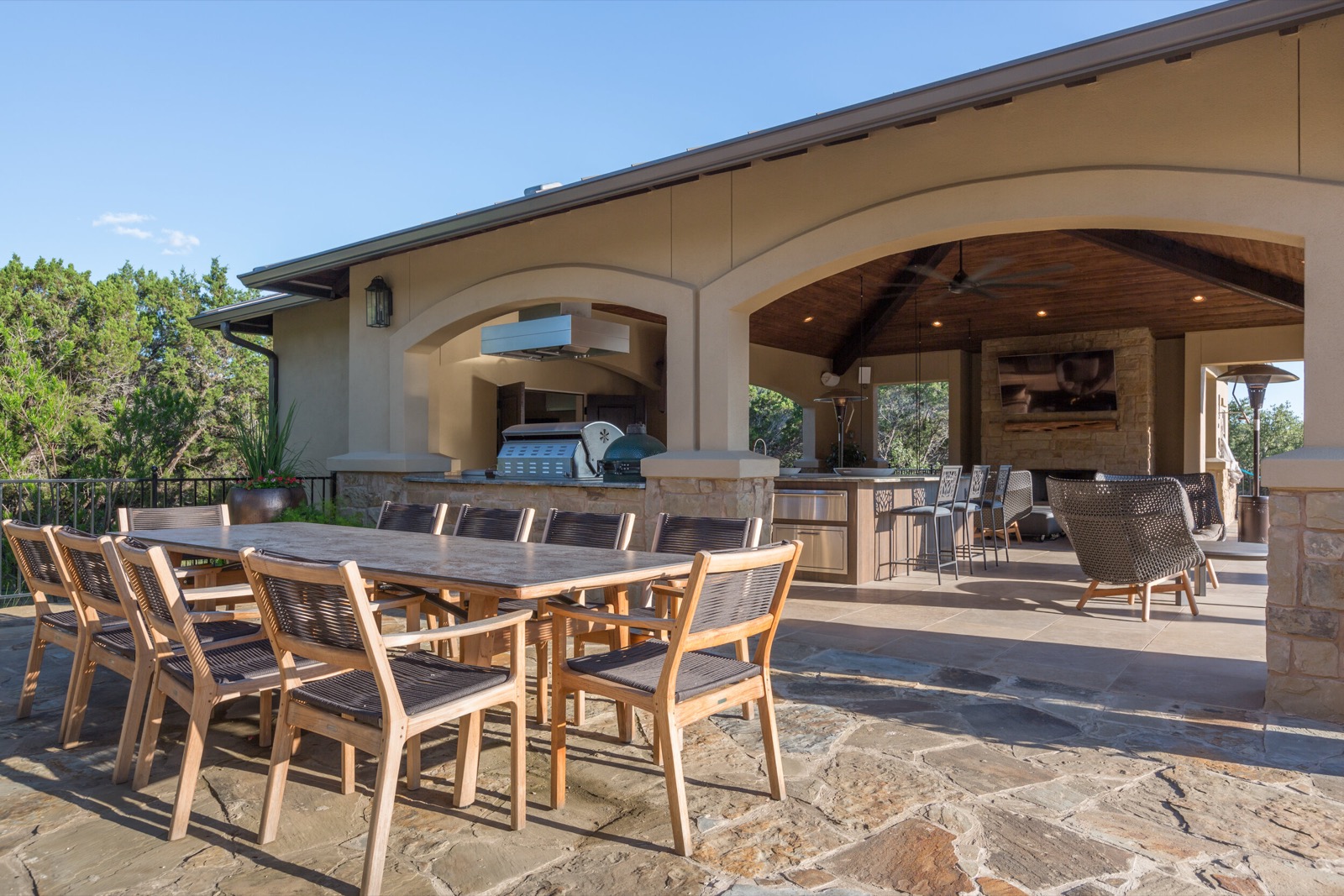 The cabana at the Lake Travis estate with its outdoor kitchen, bar, dining table, and the TV over the stone fireplace, with the hill country beyond