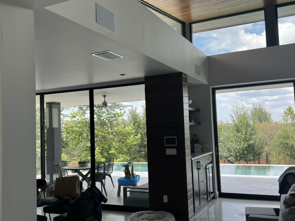 Finished living room opening onto a pool, with two discreet Triad speaker grilles near the ceiling and a Control4 touchscreen on the central dark pillar.